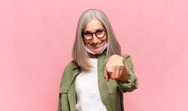 Orta yaşlı bir kadın kameraya bakıyor, kendinden emin, arkadaş canlısı bir gülümsemeyle, seni seçiyor.