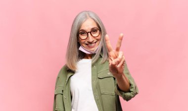 Orta yaşlı kadın gülümsüyor ve mutlu görünüyor, kaygısız ve pozitif, zafer ya da barış işareti yapıyor.