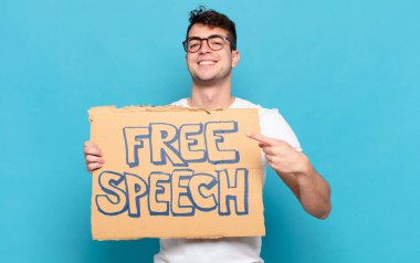 Genç adam neşeyle gülümsüyor, mutlu hissediyor ve kenarı ve yukarıyı işaret ediyor, fotokopi uzayındaki nesneleri gösteriyor.