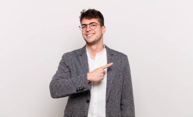 Genç adam neşeyle gülümsüyor, mutlu hissediyor ve kenarı ve yukarıyı işaret ediyor, fotokopi uzayındaki nesneleri gösteriyor.