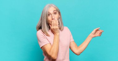 Orta yaşlı kadın kendini mutlu, şaşkın ve şaşırmış hissediyor. Ağzını eliyle kapatıyor ve yanal fotokopi alanını işaret ediyor.