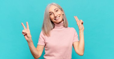 Orta yaşlı kadın gülümsüyor ve mutlu görünüyor, arkadaş canlısı ve memnun, zafer ya da barış işareti yapıyor iki eliyle