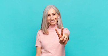 Orta yaşlı bir kadın gülümsüyor ve arkadaş canlısı görünüyor. İki ya da iki numarayı gösteriyor. Elleri önde, geriye doğru sayıyor.