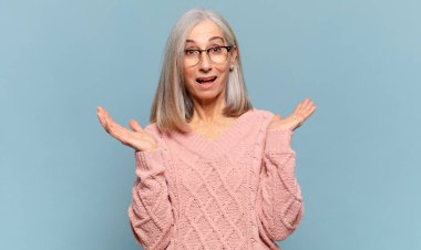 Orta yaşlı bir kadın mutlu ve heyecanlı görünüyor. Beklenmedik bir sürprizle şoke olmuş. İki eli yüzü açık.