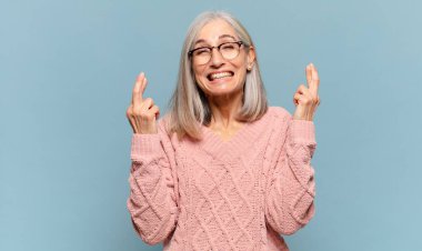 Orta yaşlı kadın gülümsüyor ve endişeyle iki parmağını çapraz yapıyor. Endişeli hissediyor, iyi şanslar diliyor ya da umut ediyor.