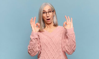 Orta yaşlı kadın şok olmuş, şaşırmış ve şaşırmış hissediyor. Onaylıyor. İki eliyle de onay işareti yapıyor.