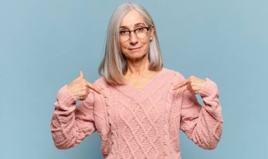 Orta yaşlı kadın gururlu, pozitif ve rahat görünüyor. İki eliyle göğsünü gösteriyor.