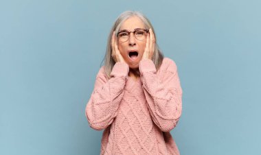 Orta yaşlı kadın mutsuz, korkmuş ya da endişeli görünüyor, ağzı sonuna kadar açık ve iki kulağını da elleriyle kapatıyor.