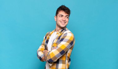 Genç adam neşeyle gülümsüyor, mutlu, tatmin olmuş ve rahatlamış hissediyor, kollarını kavuşturup yan tarafa bakıyor.