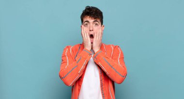 Genç adam şok olmuş ve korkmuş hissediyor, ağzı açık ve elleri yanaklarında korkmuş görünüyor.