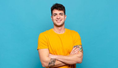 Genç adam kameraya çapraz kollarla gülümsüyor ve mutlu, kendinden emin, tatmin olmuş bir ifade, yan bakış.
