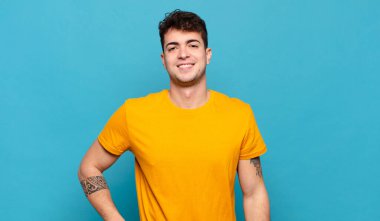 Genç adam mutlu bir şekilde gülümsüyor, eli kalçasında ve kendinden emin, olumlu, gururlu ve arkadaş canlısı.