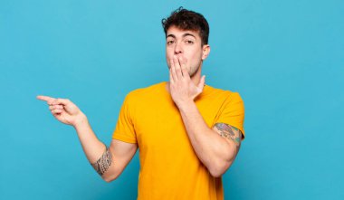 Genç adam mutlu, şaşkın ve şaşırmış hissediyor. Ağzını eliyle kapatıyor ve yanal fotokopi alanını işaret ediyor.