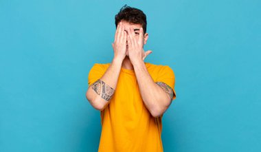 Genç adam elleriyle yüzünü kapatıyor, parmaklarının arasından şaşkın bir ifadeyle yan tarafa bakıyor.