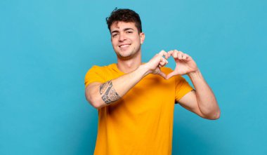 Genç adam gülümsüyor ve mutlu, sevimli, romantik ve aşık, iki eliyle kalbi şekillendiriyor.