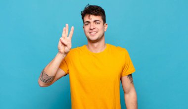 Genç adam gülümsüyor ve arkadaş canlısı görünüyor, üç ya da üç numarayı gösteriyor, eli önde, geriye doğru sayıyor.