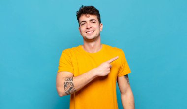 Genç adam neşeyle gülümsüyor, mutlu hissediyor ve kenarı ve yukarıyı işaret ediyor, fotokopi uzayındaki nesneleri gösteriyor.