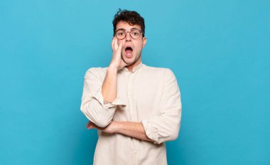 Genç adam şok ve inançsızlık içinde, elleri yanak ve kolları çapraz, sersemlemiş ve şaşırmış hissediyor.