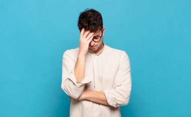 Genç adam stresli, utanmış ya da üzgün görünüyor, baş ağrısı var, yüzü elle kapatılmış.