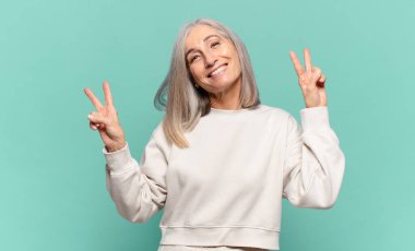 Orta yaşlı kadın gülümsüyor ve mutlu görünüyor, arkadaş canlısı ve memnun, zafer ya da barış işareti yapıyor iki eliyle