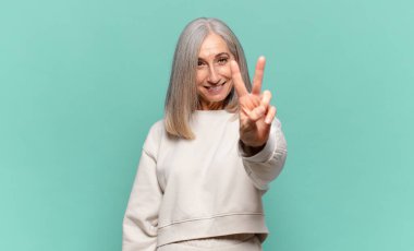 Orta yaşlı kadın gülümsüyor ve mutlu görünüyor, kaygısız ve pozitif, zafer ya da barış işareti yapıyor.