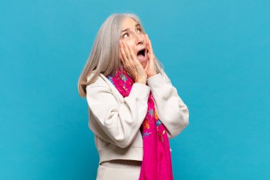 Orta yaşlı bir kadın, mutlu, heyecanlı ve şaşırmış hissediyor, iki eli yüzü yana bakıyor.