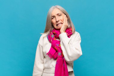 Orta yaşlı bir kadın yanağını tutuyor ve ağrılı diş ağrısı çekiyor. Hasta, sefil ve mutsuz hissediyor. Dişçi arıyor.