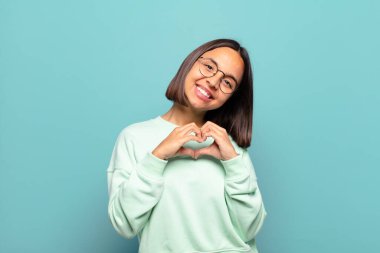 Genç İspanyol kadın gülümsüyor ve mutlu, sevimli, romantik ve aşık, iki eliyle kalbi şekillendiriyor.