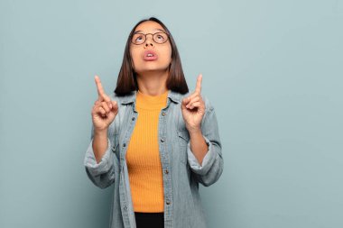 Genç İspanyol kadın şaşırmış, şaşırmış ve ağzı açık görünüyor, iki eliyle boşluğu kopyalamak için yukarı bakıyor.