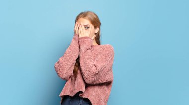 Sarışın, güzel bir kadın korkmuş ya da utanmış hissediyor. Gözleri yarı kapalı ya da ellerle dikizliyor.