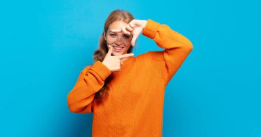 Sarışın, güzel bir kadın mutlu, arkadaş canlısı ve pozitif hissediyor, gülümsüyor ve elleriyle bir portre veya fotoğraf çerçevesi yapıyor.