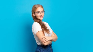 Sarışın, güzel bir kadın kameraya çapraz kollarıyla gülümsüyor ve mutlu, kendinden emin, tatmin olmuş bir yüz ifadesi var.