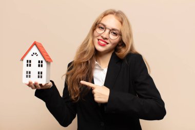 Sarışın kadın neşeyle gülümsüyor, mutlu hissediyor ve yan ve yukarı doğru işaret ediyor, fotokopi uzayındaki nesneleri gösteriyor.