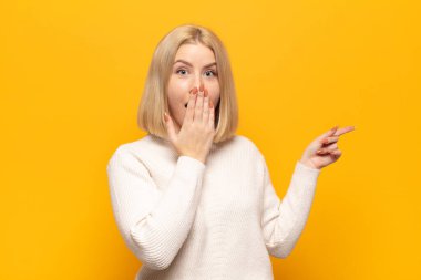 Sarışın kadın mutlu, şaşkın ve şaşırmış hissediyor. Ağzını eliyle kapatıyor ve yanal fotokopi alanını işaret ediyor.