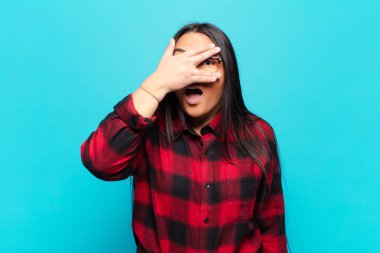 Şok olmuş, korkmuş ya da korkmuş görünen Latin bir kadın. Yüzü elle örtülmüş ve parmaklarının arasından gizlice bakıyor.