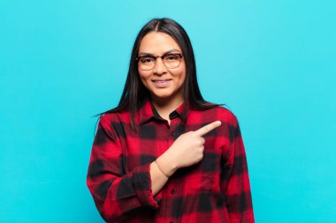 Latin kadın neşeyle gülümsüyor, mutlu hissediyor ve yan ve yukarı doğru işaret ediyor, kopyalama alanında nesne gösteriyor