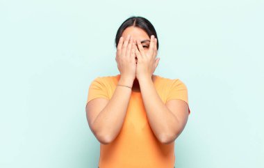 Latin kadın elleriyle yüzünü kapatıyor, parmaklarının arasından şaşkın bir ifadeyle yan tarafa bakıyor.