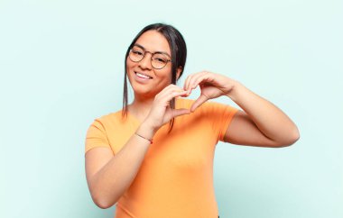 Latin kadın gülümsüyor ve mutlu, sevimli, romantik ve aşık, iki eliyle kalbi şekillendiriyor.