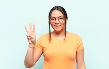 Latin kadın gülümsüyor ve arkadaş canlısı görünüyor. Üç ya da üç numarayı gösteriyor. Elleri önde, geriye doğru sayıyor.