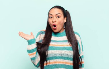 Latin kadın şaşırmış ve şoka uğramış görünüyor, çenesi düşmüş ve eli açık bir objeyi tutuyor.