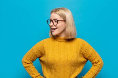 Sarışın kadın mutlu, neşeli ve kendinden emin görünüyor. Gururla gülümsüyor ve iki elini de kalçasında istiyor.