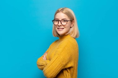 Sarışın kadın kameraya çapraz kollarıyla gülümsüyor ve mutlu, kendinden emin, tatmin olmuş bir ifade var.