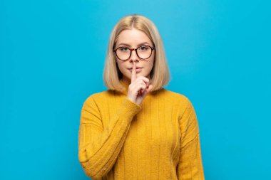 Sarışın kadın ciddi görünüyor ve parmaklarını dudaklarına bastırıp sessizlik ya da sessizlik talep ediyor, sır saklıyor.