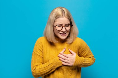 Sarışın kadın üzgün, incinmiş ve kalbi kırık görünüyor, iki elini de kalbe yakın tutuyor, ağlıyor ve depresyonda.