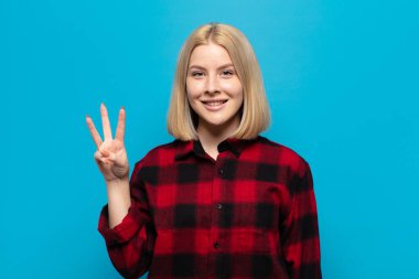 Sarışın kadın gülümsüyor ve arkadaş canlısı görünüyor, üç ya da üç numarayı gösteriyor, eli önde, geriye doğru sayıyor.