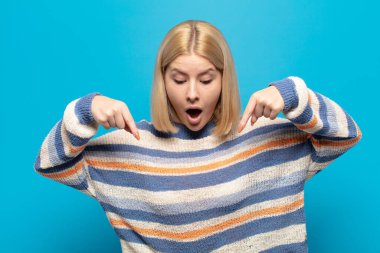 Sarışın kadın şaşkın, ağzı açık ve şaşkın, inançsızlık ve şaşkınlık içinde bakıyor ve onu işaret ediyor.