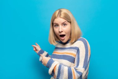 Sarışın kadın şaşkın ve şaşırmış hissediyor. Yan taraftaki fotokopi alanını işaret ediyor. Şaşırmış, ağzı açık bir şekilde bakıyor.