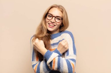 Sarışın kadın sevinçle gülümsüyor ve kutluyor, yumruklarını sıkmış ve kollarını kavuşturmuş, mutlu ve pozitif hissediyor.