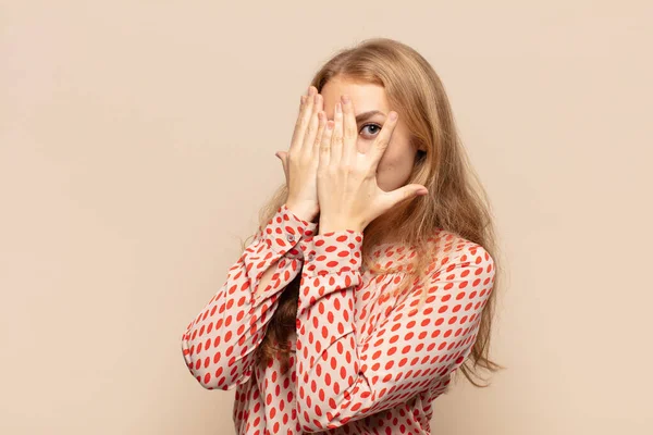 blonde woman covering face with hands, peeking between fingers with surprised expression and looking to the side