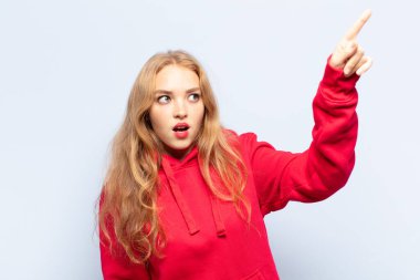 Şok olmuş ve şaşırmış hisseden sarışın bir kadın, şaşkınlık içinde, ağzı açık bir bakışla yukarı bakıyor ve onu işaret ediyor.